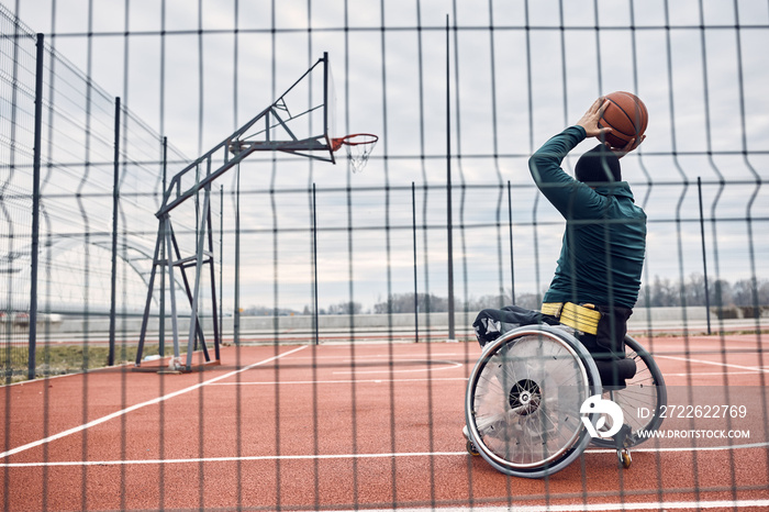 Rear view of wheelchair-bound basketball player taking a shot while practicing on the court outdoors.