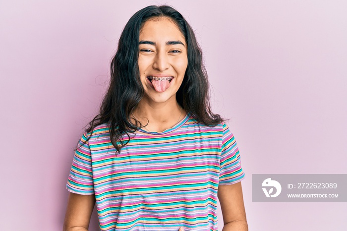 Hispanic teenager girl with dental braces wearing casual clothes sticking tongue out happy with funny expression. emotion concept.