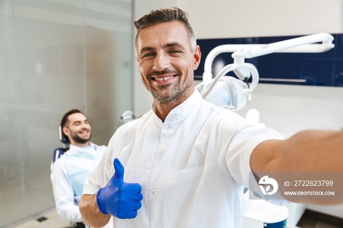 Handsome happy young man doctor in medical dentist center showing thumbs up take a selfie by camera.