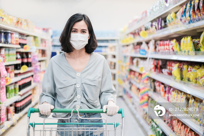 Asian woman wearing face mask and rubber glove push shopping cart in suppermarket departmentstore. Girl choosing, looking grocery things to buy at shelf during coronavirus crisis or covid19 outbreak.