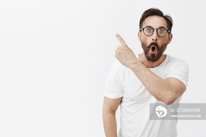 Fantastic, look. Portrait of amazed and impressed handsome adult man with beard and wrinkles in glasses, pointing at upper left corner, dropping jaw and sharing impressions with friend