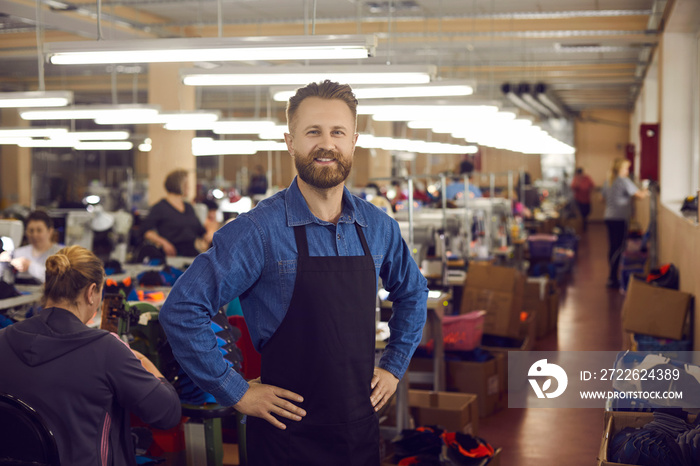 shoemaker. Portrait of a confident smiling factory worker or shoe factory owner at work. Caucasian handsome bearded man in a black work apron holds his hands to his sides looking at the camera.