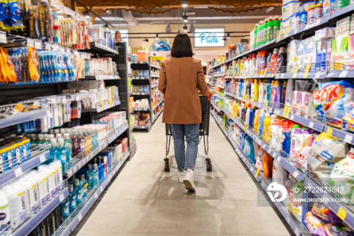 a woman with a cart walks between rows of shelves in a grocery store