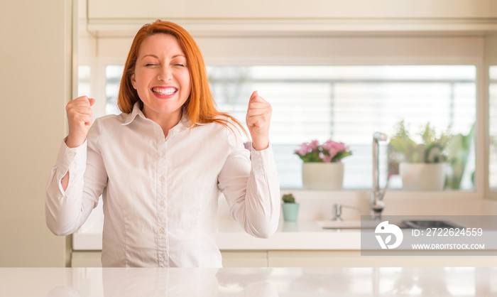 Redhead woman at kitchen excited for success with arms raised celebrating victory smiling. Winner concept.