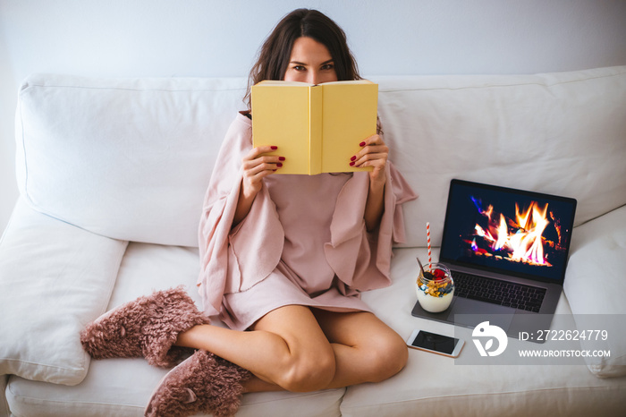 Girl hiding her face behind a book while sitting on the sofa with a fireplace on the laptop - young woman relaxing and reading in cozy room - hardcover book mockup