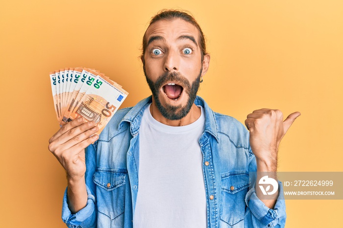 Attractive man with long hair and beard holding bunch of 50 euro banknotes pointing thumb up to the side smiling happy with open mouth