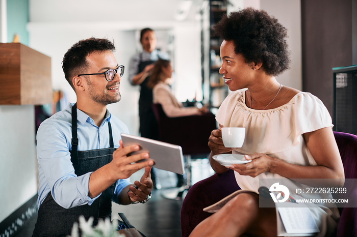 Happy hairdresser and African American woman use digital tablet while talking at hair salon.