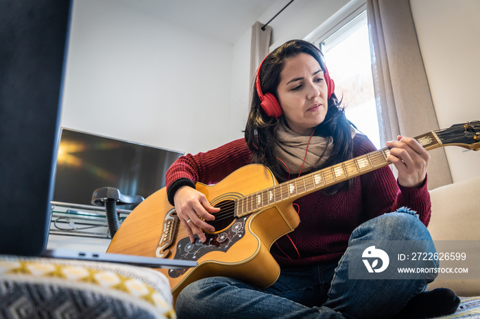 Stock photo of concentrated woman playing her guitar and singing while following tutorial in her laptop.