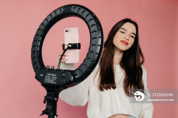 Attractive young lady is smiling and looking at the camera. The girl is adjusting a mobile phone to the ring light. Young woman is taking selfies for the fashion blog. Concept of photography