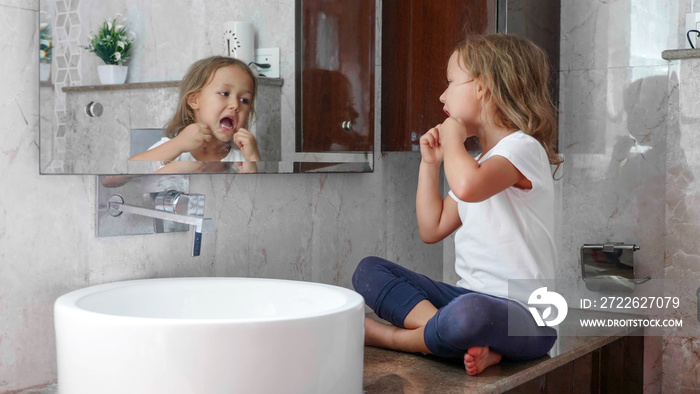 Little cute girl brushes her teeth with dental floss while sits on the table near the sink at domestic bathroom in front of the mirror.