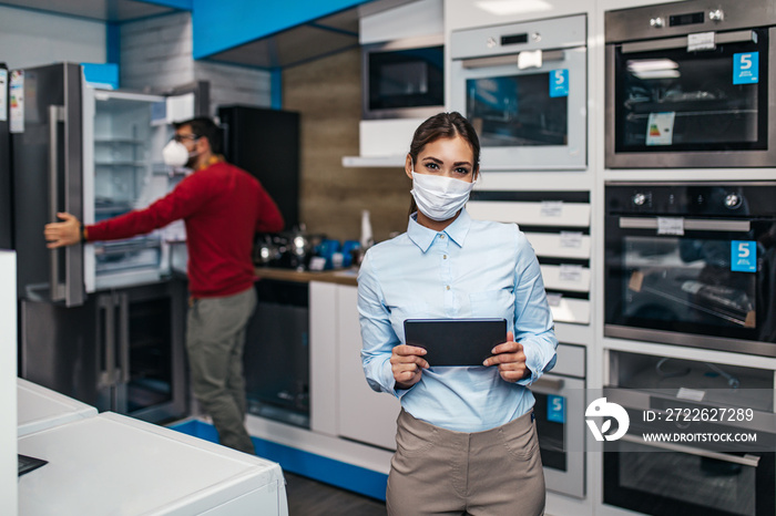 Young saleswoman working in store with household appliances. She is wearing face protective mask and holding tablet. Pandemia, Covid-19 concept.