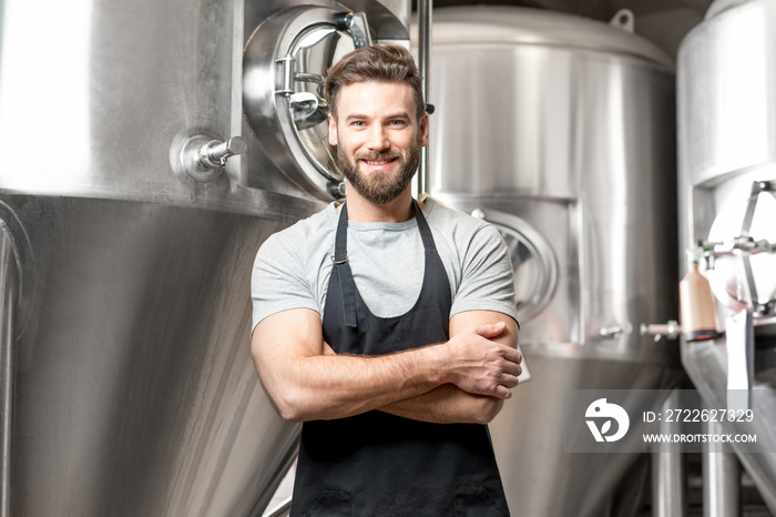 A portrait of handsome brewer in uniform at the beer manufacture with metal containers on the background