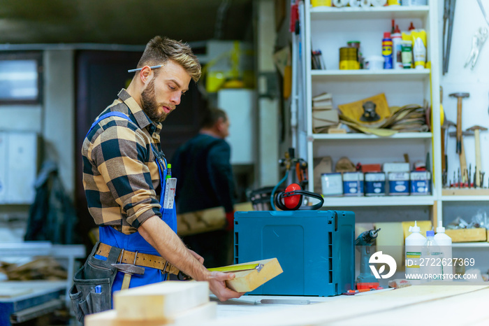 Portrait of young handsome caucasian carpenter who measures a board at the workshop