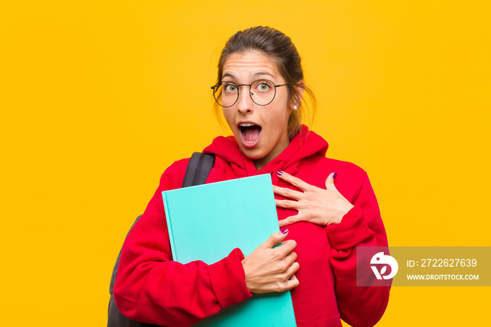 young pretty student feeling shocked, astonished and surprised, with hand on chest and open mouth, saying who, me?