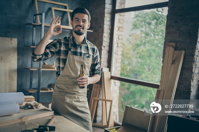 Photo of cheerful bearded positive man showing ok sign in apron checkered shirt holding bottle of beer having finished woodwork