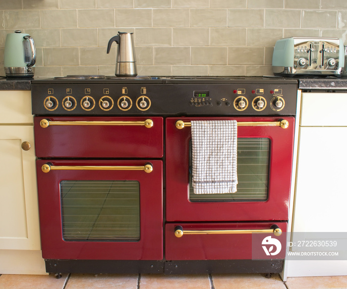 Red Range style stove cooker with a silver mocha coffee pot on a hot plate. Good symmetry and natural light. A white tea towel hangs on a gold oven rail. Simple image with a minimalist coffee theme.