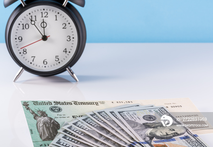 Stack of 100 dollar bills with illustrative coronavirus stimulus payment check and alarm clock to show delay in receiving money