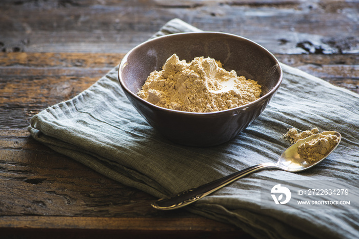 Maca Pulver in einer Keramikschüssel auf einem rustikalen Holztisch mit Stoffunterlage und Silberlöffel - peruanisches Maca Powder - lat. Lepidium meyenii - Peruanischer Ginseng