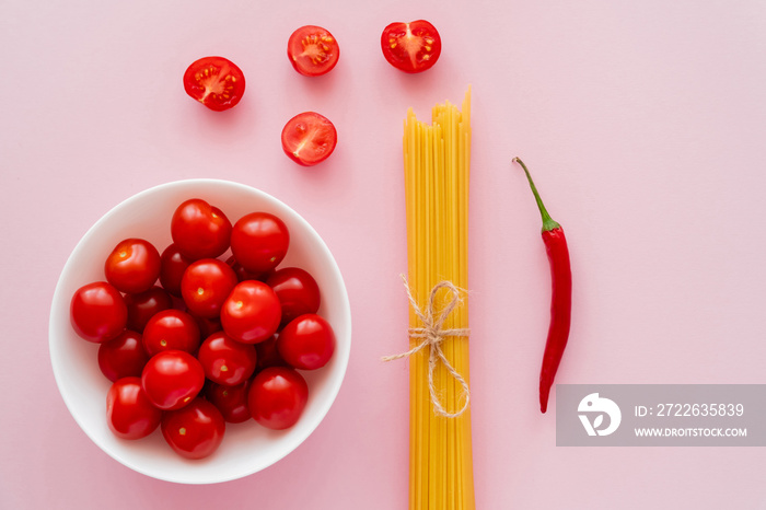 Top view of cherry tomatoes in bowl near raw pasta and chili pepper on pink surface.