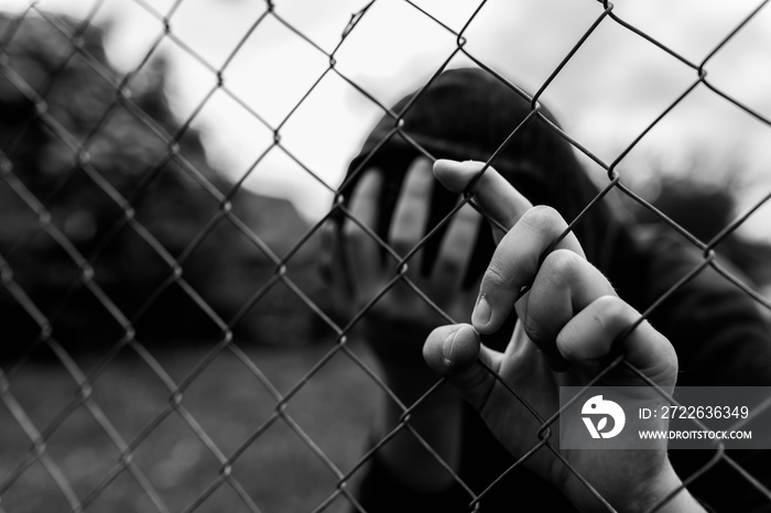 Young unidentifiable teenage boy holding the wired garden at the correctional institute , covering hes face in black and white, conceptual image of juvenile delinquency, focus on the boys hand.