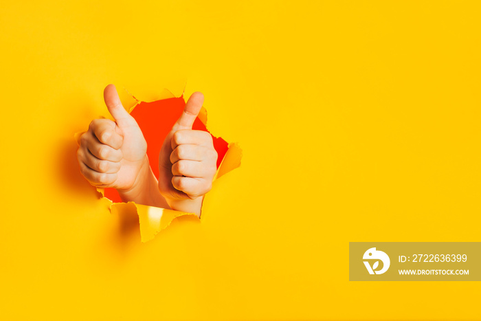 Two hands show OK gesture, approval, liking, acceptance through torn hole in yellow paper wall. Double thumbs up sign. A side place for text information, product or service advertising. Copy space.