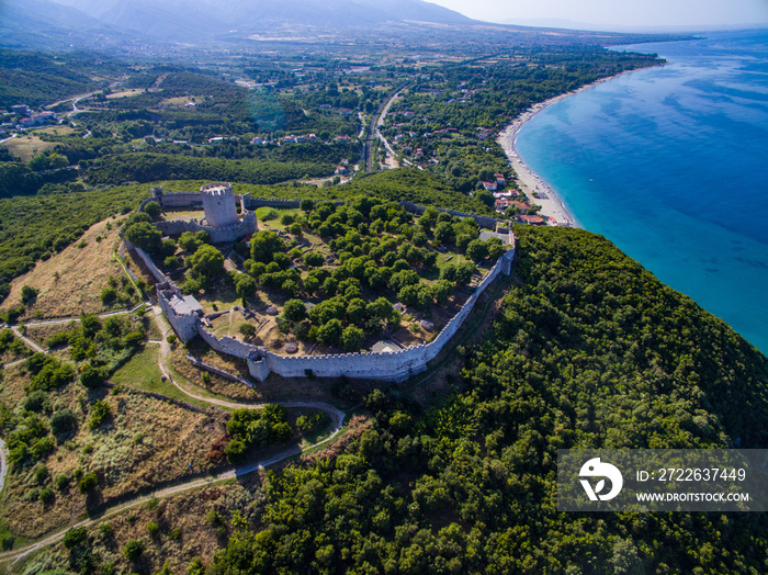 An aerial perspective of the medieval castle of platamonas on the top of a green hill looking down at the beach.