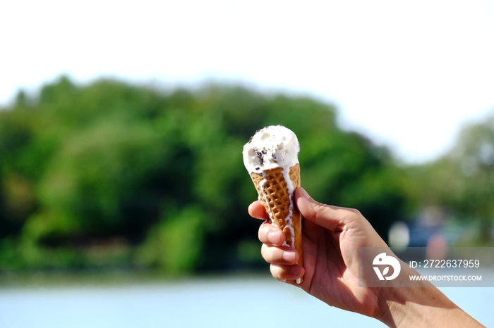 A male hand holding a soft cones of melting ice cream with blurred green nature background in sunshine day and water view