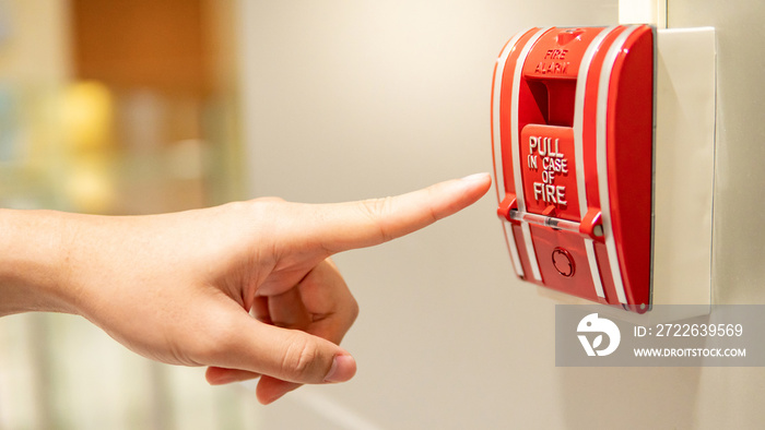 Male hand pointing at red fire alarm switch on concrete wall in office building. Industrial fire warning system equipment for emergency.