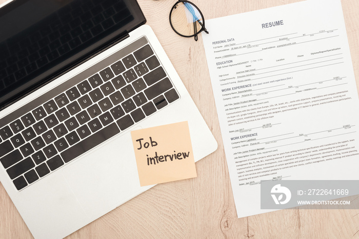 top view of laptop, resume template, glasses and sticky note with job interview lettering on wooden table