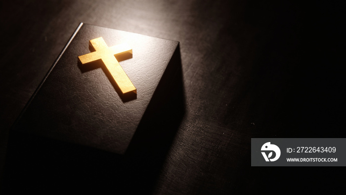 Golden Christian cross resting on Bible illuminated by light coming from above.