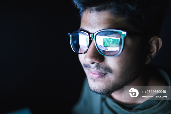 Hacker in glasses and a hood works at a computer in the dark, a reflection in glasses close-up.