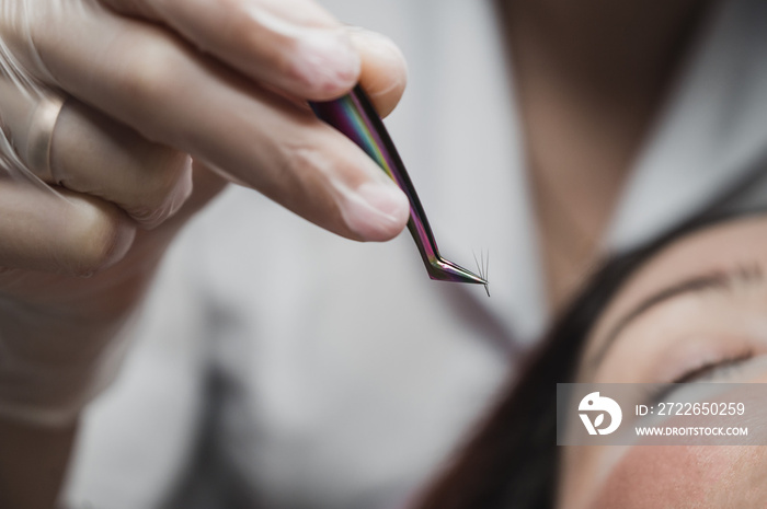 Woman lies on eyelash extension procedure in a beauty salon. Lashmaker holds tweezers with a bunch of artificial eyelashes. Close-up of a craftsman in gloves.