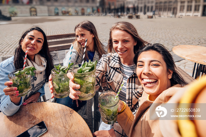 group of multiethnic young happy cheerful women sitting on terrace of the city center with cocktails in their hands taking a selfie