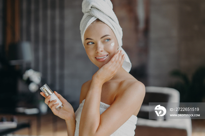 Indoor shot of young smiling woman applies moisturizer cream on face, takes care of her skin and complexion, puts lotions, has minimal makeup, wrapped in bath towel. Beauty, cosmetology concept