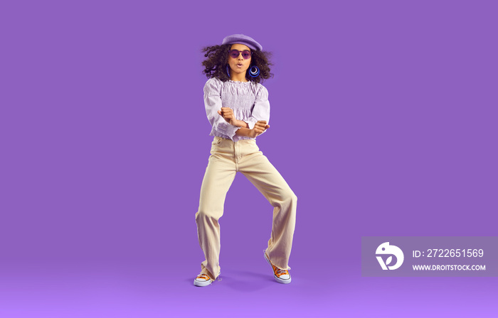 Child dancing. Confident African American girl with curly hair in trendy top, beige jeans, beret hat and sunglasses dancing gangnam style on purple studio background. Full body. Kids’ fashion concept