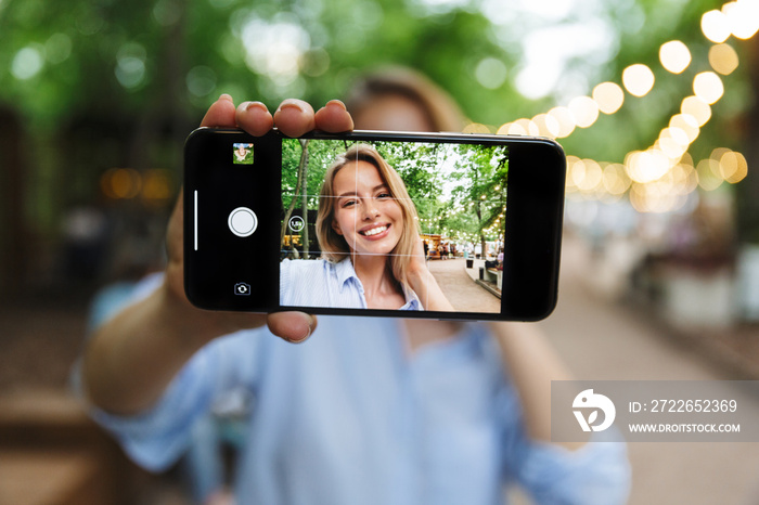 Excited happy young woman posing outdoors in park take a selfie by mobile phone.