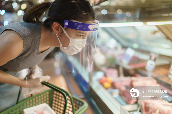 Woman wearing gloves, face shield and mask choosing meat at supermarket . Panic shopping during the corona virus pandemic.