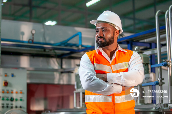 Professional engineer with helmet works to maintain industrial construction equipment. Worker is standing in the factory with their arms crossed.