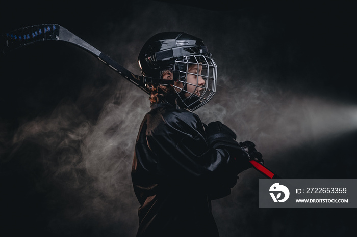 Young blonde sporty boy, ice hockey player, posing in a dark studio for a photoshoot, wearing an ice-skating uniform, helmet, gloves and holding up a hockey stick, confident look from the side