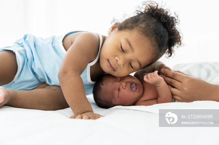 African American little girl kissing on newborn baby cheek on white bed at home. Little girl takes care of infant baby with kindly