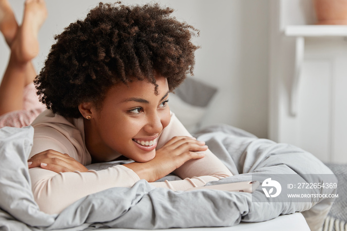 Close up shot of relaxed beautiful happy dark skinned young female with curly hair, lies in comfortable bed, being in good mood after unforgettable night with husband, enjoys day off or weekend