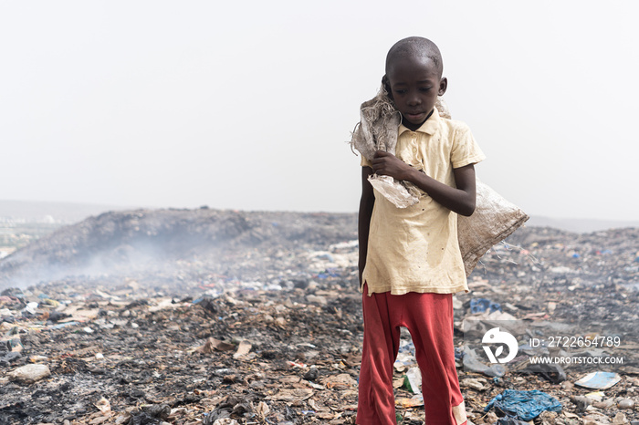 Neglected, malnourished African boy standing desolate in the midst of a smelly and smoking garbage dump; social issue of child abandon