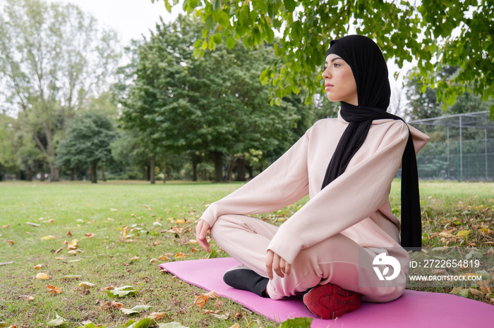 Woman in hijab and pink tracksuit meditating in park