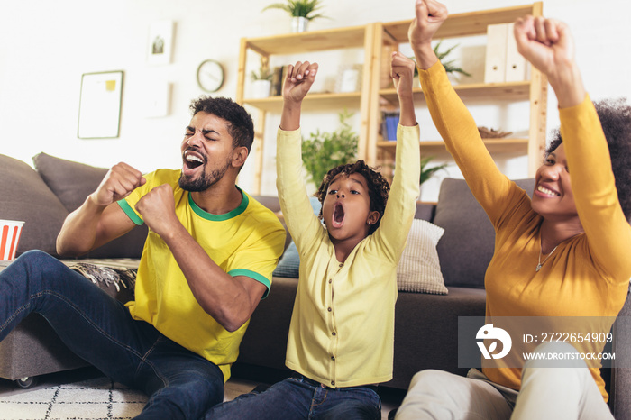 Happy African American family of three watching tv and cheering sport games on sofa at home