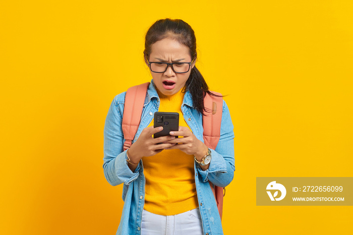 Portrait of surprised young Asian woman student in casual clothes with backpack using mobile phone, typing sms messages isolated on yellow background. Education in college university concept