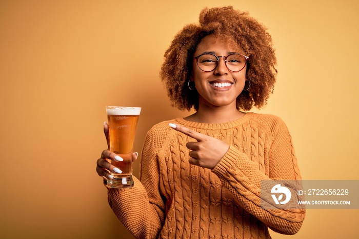 Young beautiful African American afro woman with curly hair drinking glass of beer very happy pointing with hand and finger