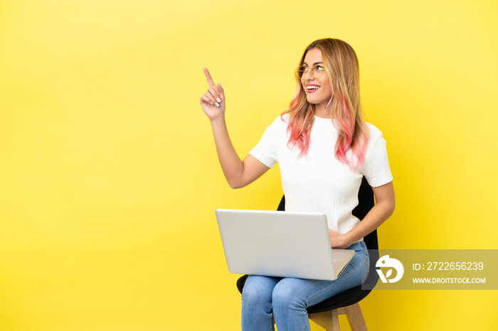 Young woman sitting on a chair with laptop over isolated yellow background pointing up a great idea