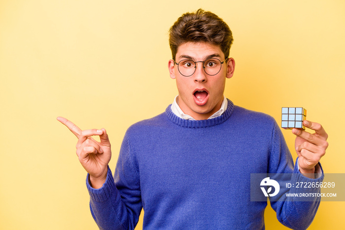 Young caucasian man holding a Rubiks cube isolated on yellow background pointing to the side