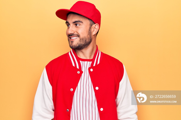 Handsome sporty man with beard wearing baseball jacket and cap over yellow background looking to side, relax profile pose with natural face and confident smile.