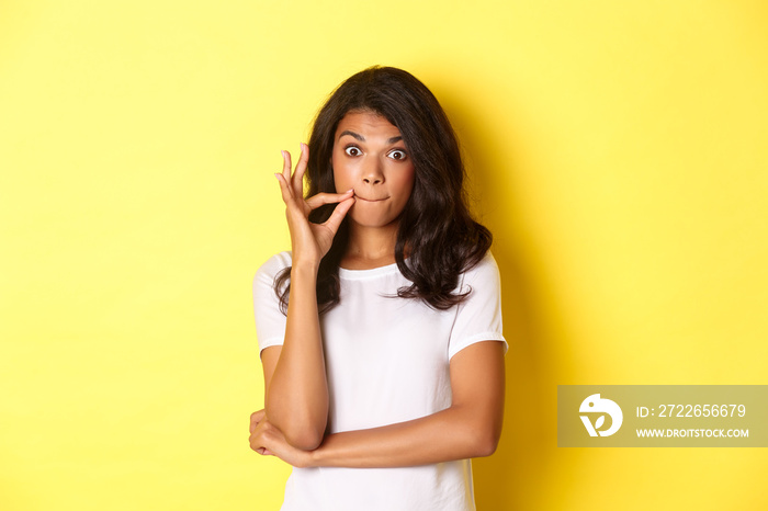 Portrait of young african-american woman making promise to keep secret, seal lips, zipping mouth with fingers, standing over yellow background